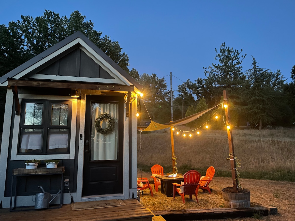 The cozy tiny home is illuminated by warm outdoor string lights, creating a welcoming ambiance. A shaded seating area features a table surrounded by four red chairs, inviting relaxation. Nearby trees provide a natural backdrop to the peaceful setting.
