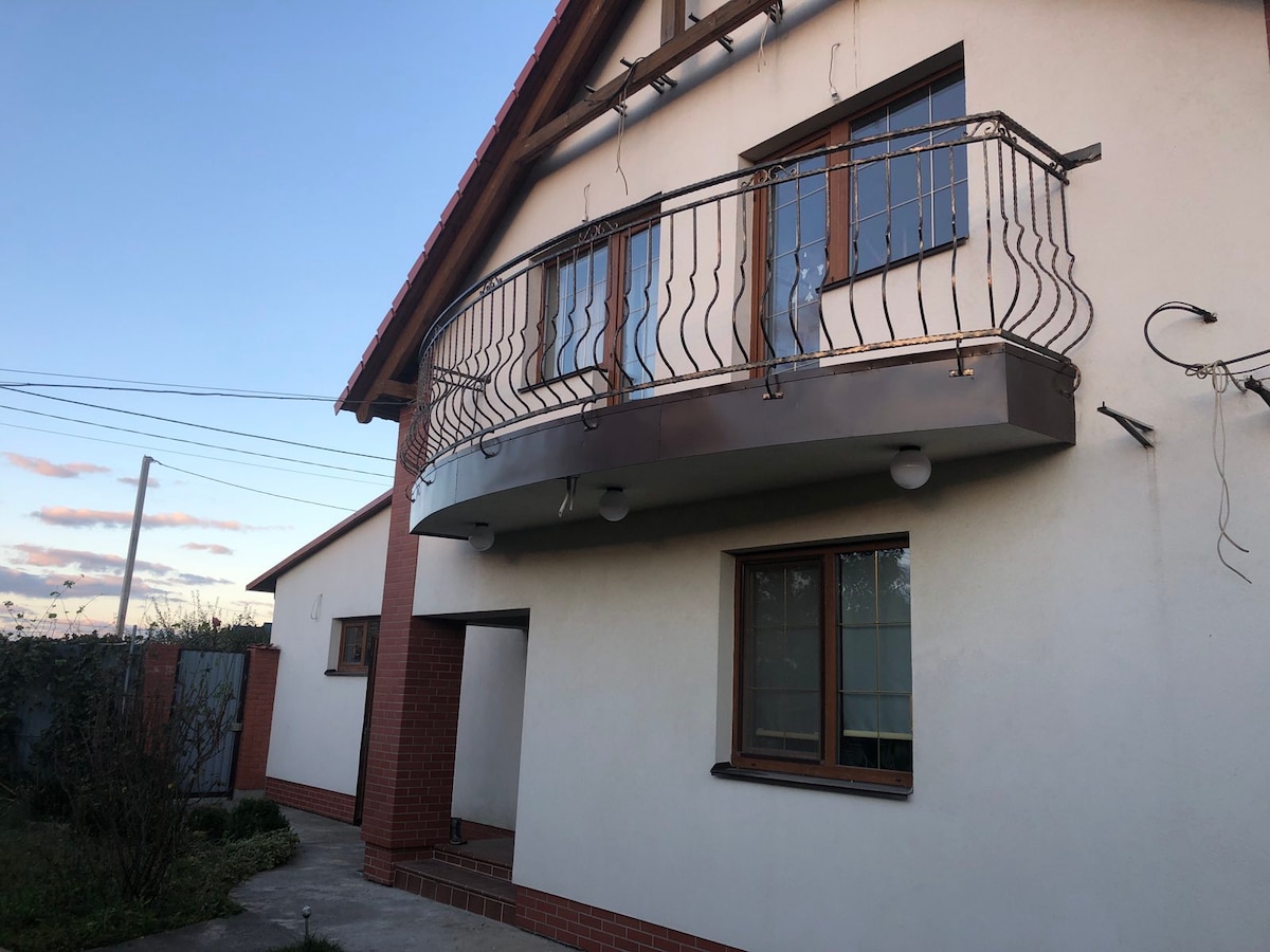 A modern two-story house is shown with a spacious balcony that features decorative railings. Large windows are present on both the upper and lower levels, allowing natural light to enter. A well-maintained garden area surrounds the entryway, contributing to a welcoming exterior.