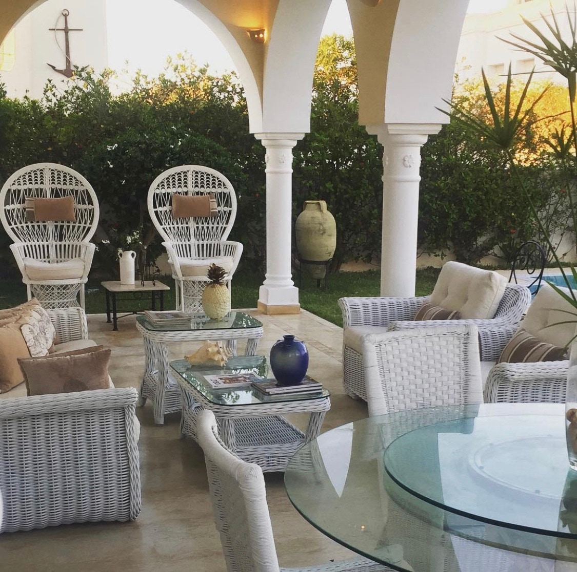A relaxed outdoor seating area is set beneath elegant arches, featuring two white wicker chairs flanking a transparent glass table. Cushions add comfort to the seating, while greenery provides a natural backdrop. Decorative items, including a vase and a terracotta pot, are arranged thoughtfully.