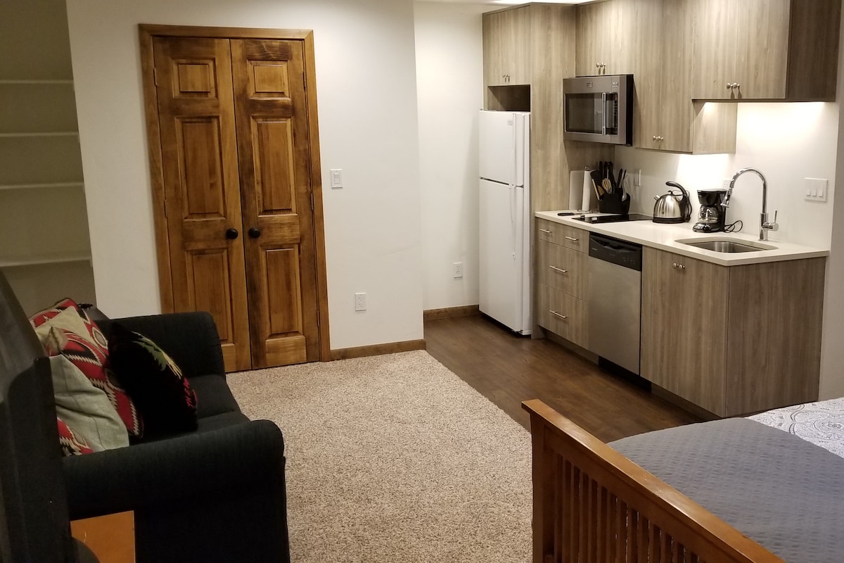 A modern studio space is presented, featuring a kitchenette with stainless steel appliances. A cozy sitting area is to the left, accompanied by a foldable table for dining or work. Light wood cabinetry and neutral tones add to the inviting atmosphere.