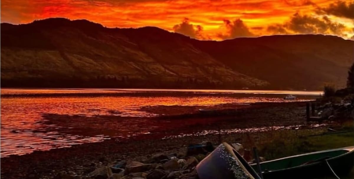 A vibrant sunset paints the sky in shades of orange and red, reflecting on the calm waters of the loch. The silhouette of a mountainous backdrop adds depth to the scene, while a rocky shoreline is visible in the foreground.