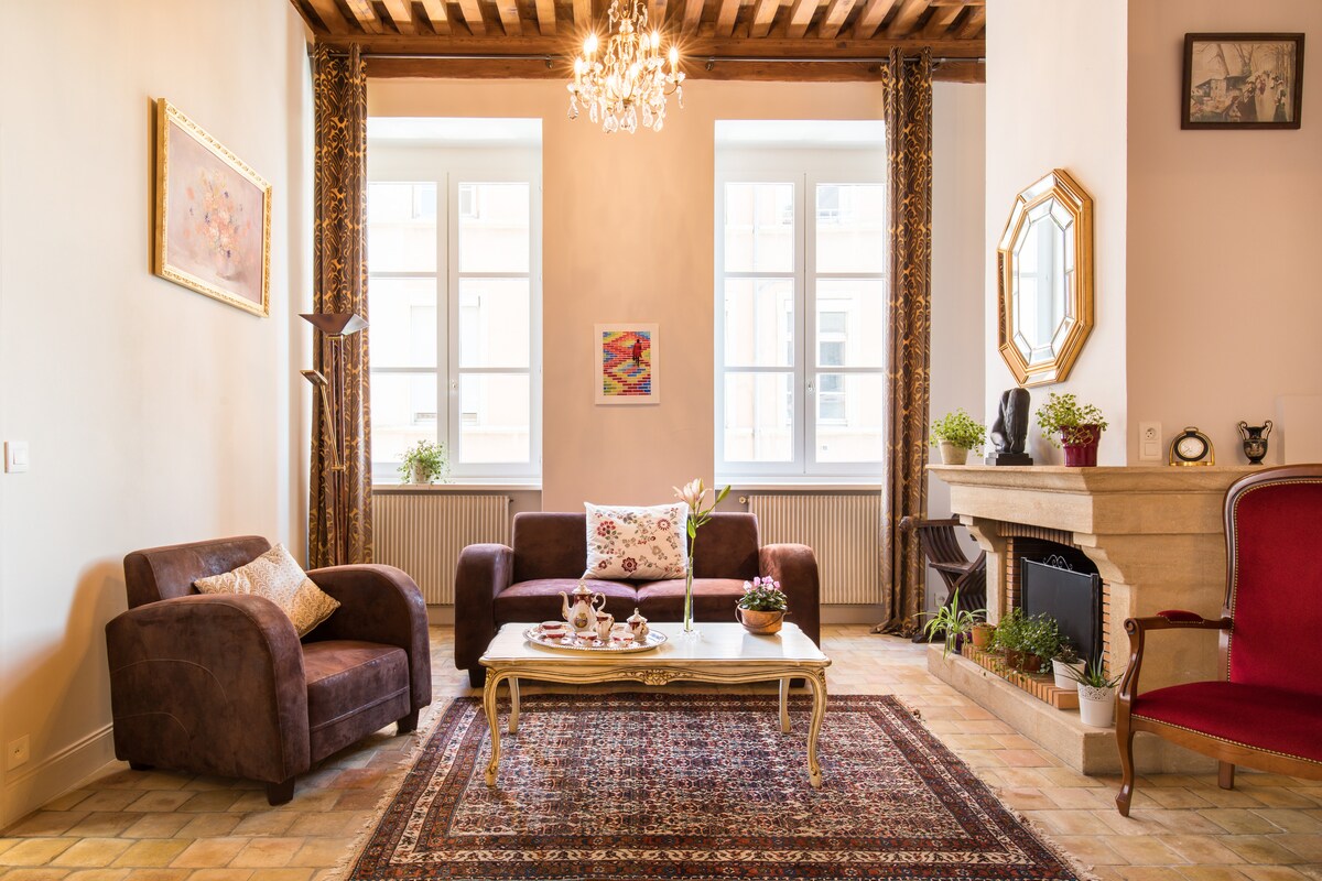 A cozy living area features two sofas and an elegant armchair, arranged around a decorative coffee table. Natural light fills the space through large windows adorned with curtains. A charming chandelier hangs from the ceiling, complementing the warm tones of the furnishings.