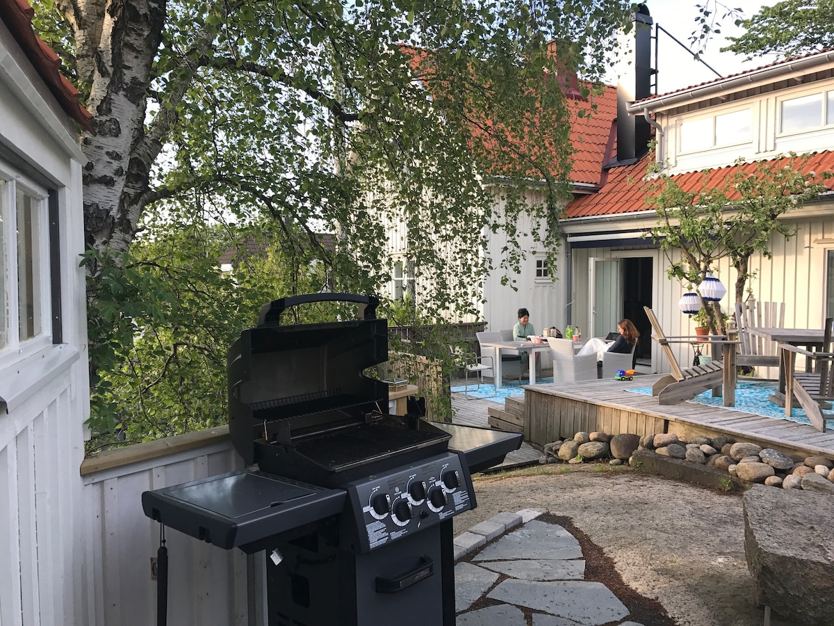 A barbecue grill is positioned in the foreground, surrounded by a stone-paved area. Visible in the background is a cozy seating area with people gathered, while the house features a blend of white and wooden exteriors, framed by lush greenery.