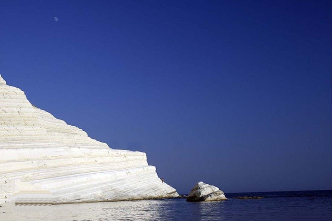 The image depicts a serene coastal landscape featuring a striking white rock formation alongside calm waters. A clear blue sky extends overhead, with a small moon visible in the upper left corner. The natural beauty of the scene highlights the tranquility of the environment.