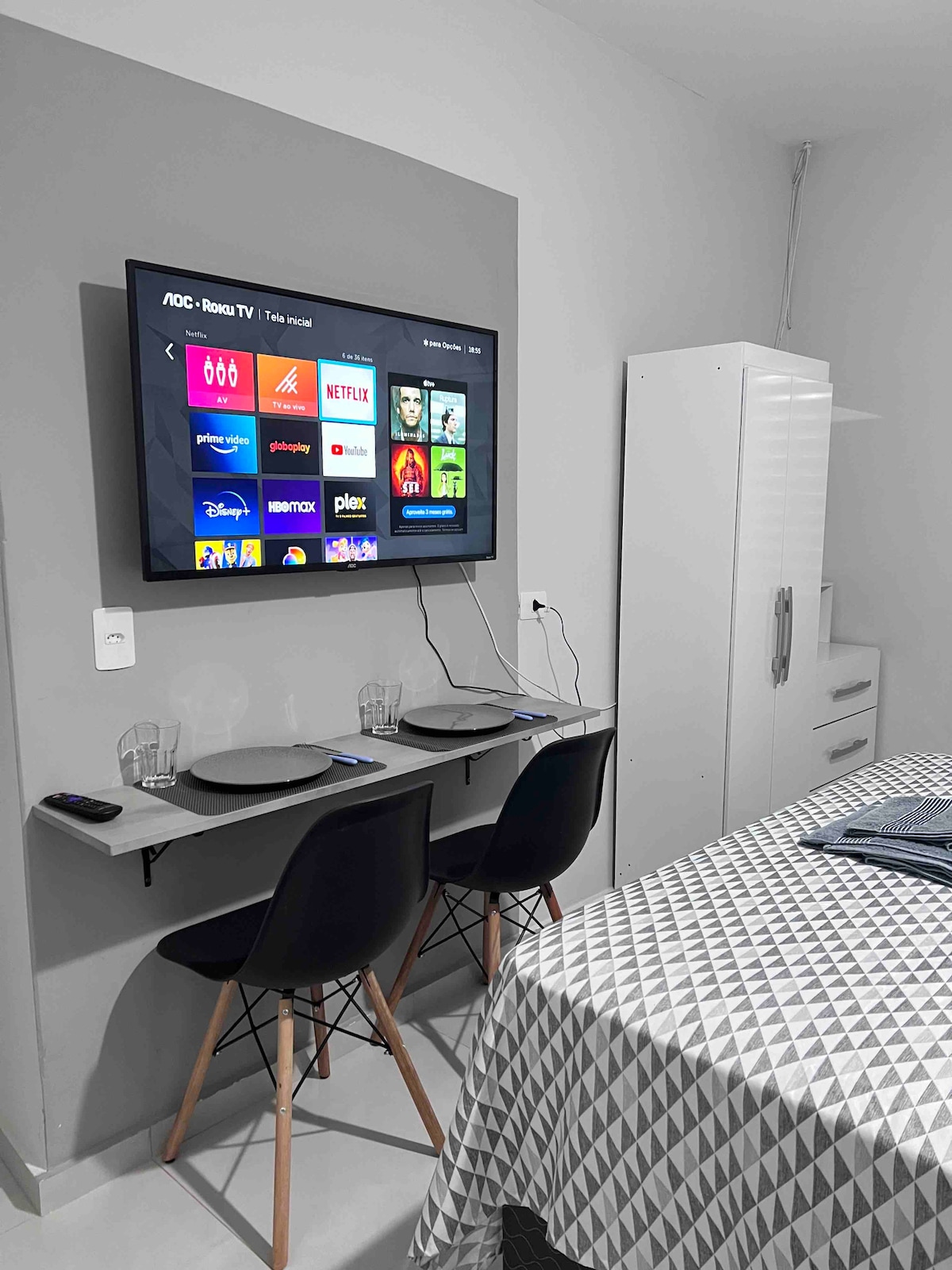 A modern living space features a Smart TV mounted on the wall displaying various streaming options. Two sleek chairs are positioned at a small table set for two, with a geometric-patterned bedspread covering a spacious bed in the background.