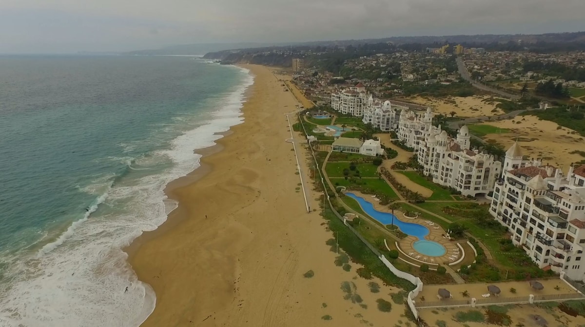 Aerial view of the coastline reveals a sprawling beach with gentle waves lapping at the shore. The elegant resort complex is surrounded by well-maintained gardens and features multiple swimming pools, with sandy stretches extending in both directions along the coastline.