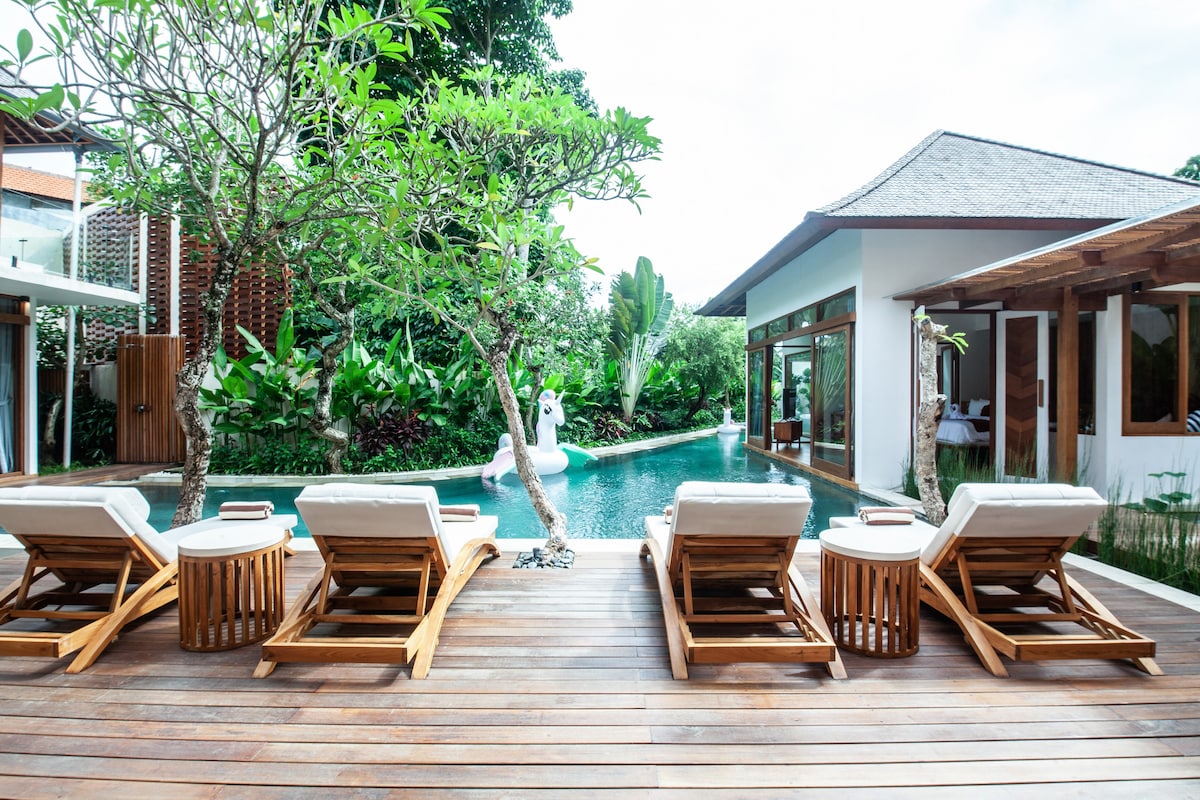 A wooden deck features four sun loungers positioned near a private infinity pool. Lush tropical greenery surrounds the area, providing a serene atmosphere. The villa's architecture is visible in the background, showcasing large glass doors that invite natural light.