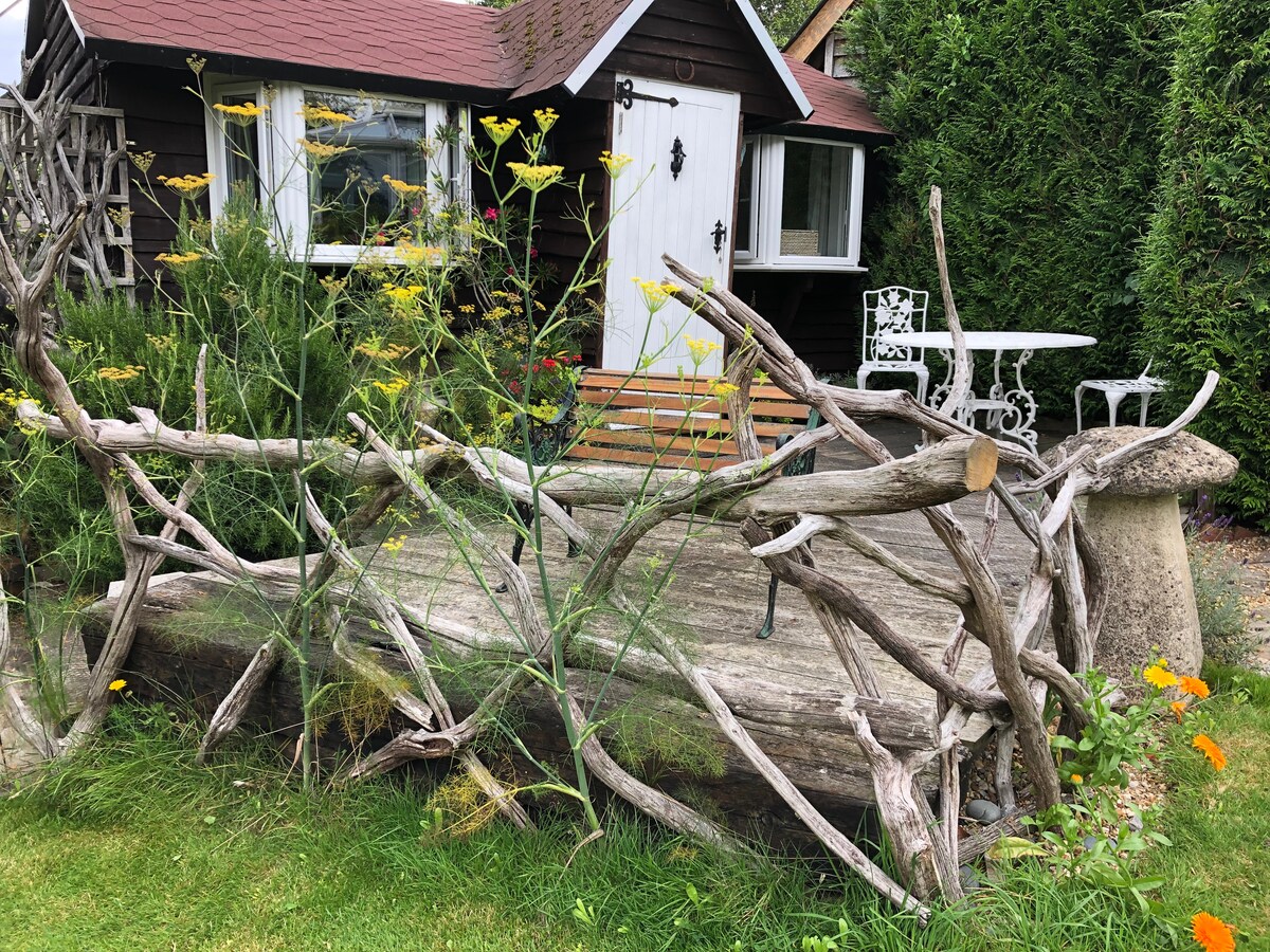A charming entrance to the garden chalet is framed by rustic wooden branches, creating a natural barrier. A small patio with a white table and chairs is visible, surrounded by lush greenery and seasonal flowers, offering a peaceful outdoor space.