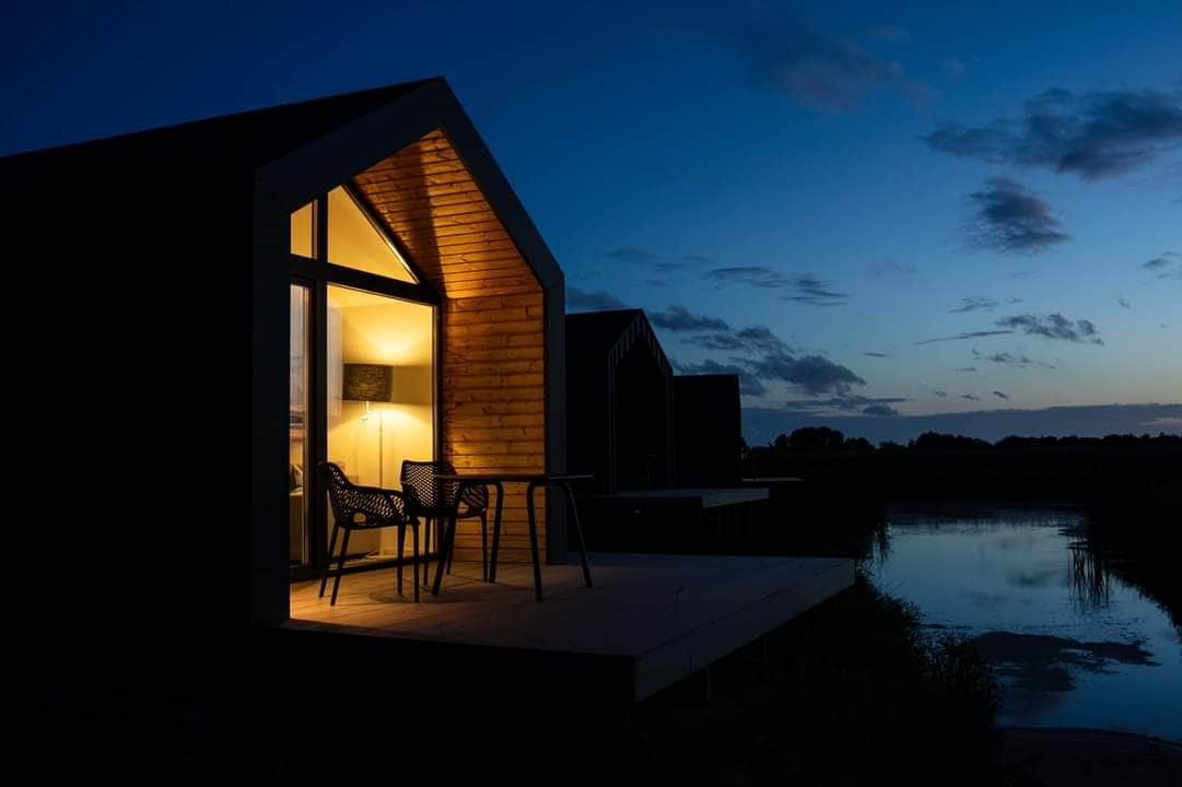 An inviting exterior of a modern vacation home is illuminated at dusk, showcasing a cozy seating arrangement on the patio. The structure features large glass doors revealing warm interior lighting, while a serene waterway reflects the evening sky.