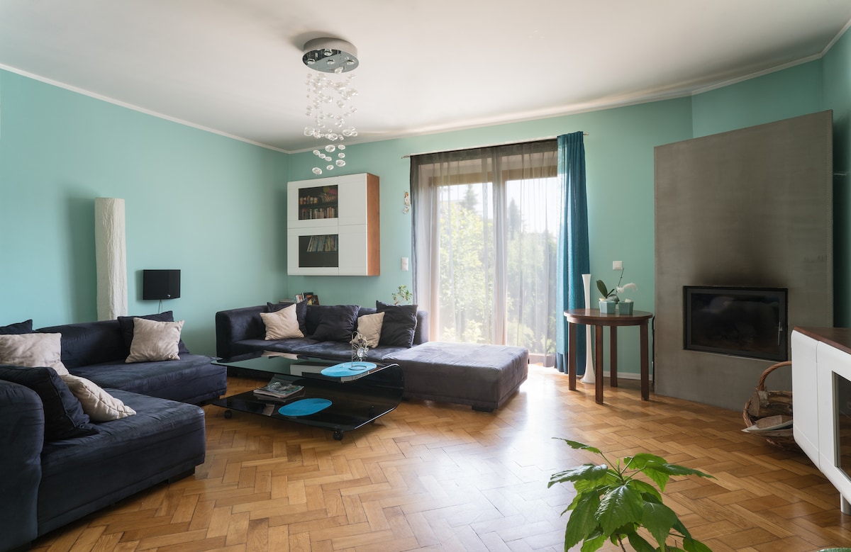A spacious living room features comfortable navy sofas arranged around a glass coffee table. Natural light fills the room through large windows, with greenery visible outside. A sleek entertainment unit is positioned against a gray wall, while a modern light fixture hangs from the ceiling.