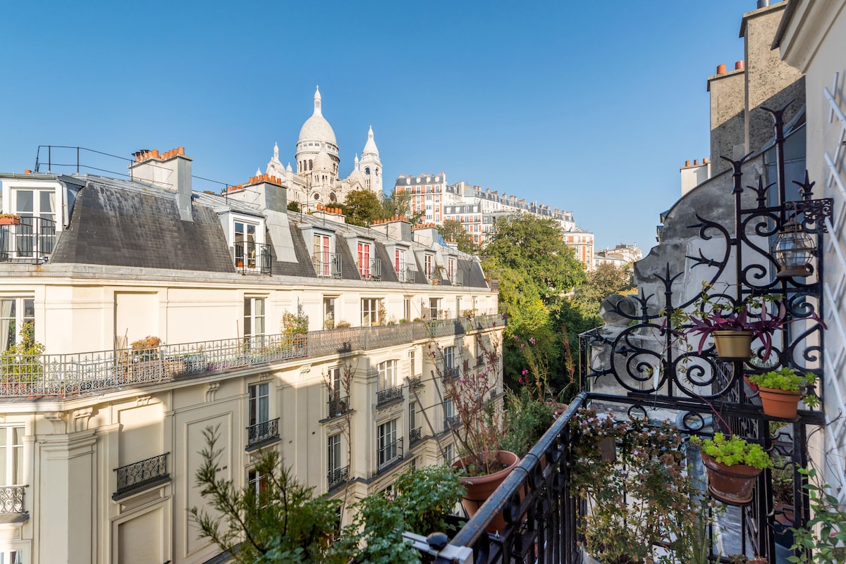 A balcony view showcases the basilica of Sacre Coeur rising majestically against a clear blue sky. Lush green plants are arranged in pots along the balcony railing, with charming Parisian rooftops visible in the background.