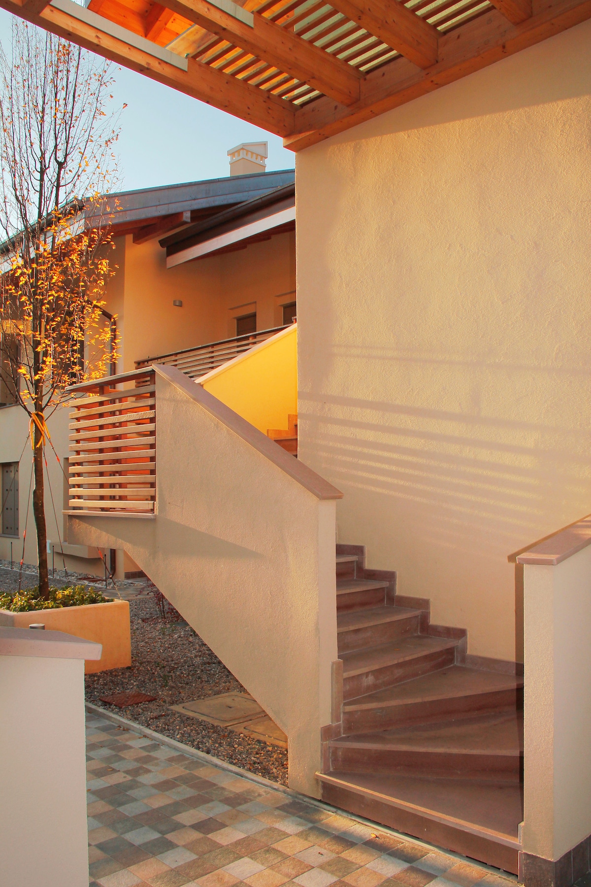 A staircase with a wooden railing leads to an upper level, casting soft shadows on the wall. The surrounding area is landscaped with small trees and gravel, while a patterned stone pathway complements the modern architecture of the adjacent building.