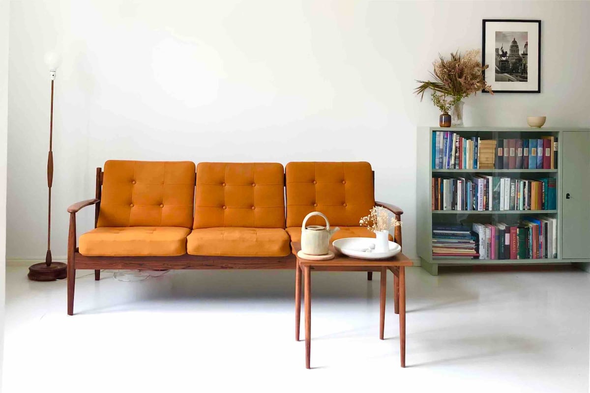 A mid-century modern sofa with three orange cushions is positioned against a white wall. A small wooden coffee table sits in front, topped with a bowl and a vase of dried flowers. A light green bookshelf is filled with an array of colorful books and a framed image is displayed above the couch.