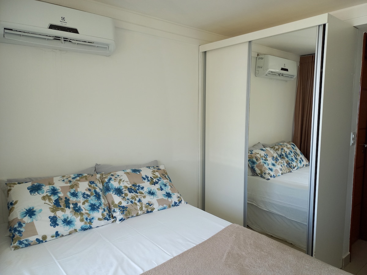 A comfortable sleeping area is featured with a spacious bed adorned with floral-patterned pillows. A wardrobe with mirrored doors reflects the room, while an air conditioning unit is mounted on the wall, ensuring a cool environment.
