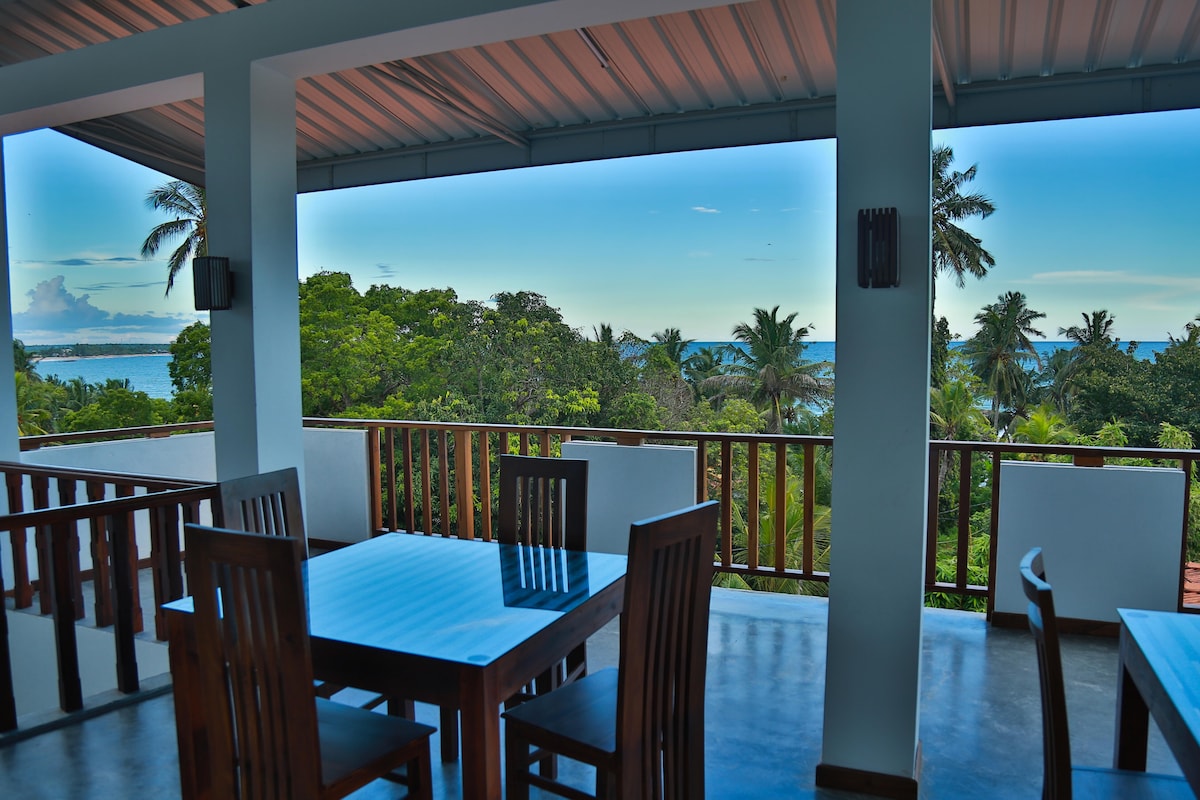 A dining area features wooden tables and chairs, set against a backdrop of lush greenery and distant ocean views. Large windows allow natural light to brighten the space, while the covered balcony creates a comfortable outdoor atmosphere.