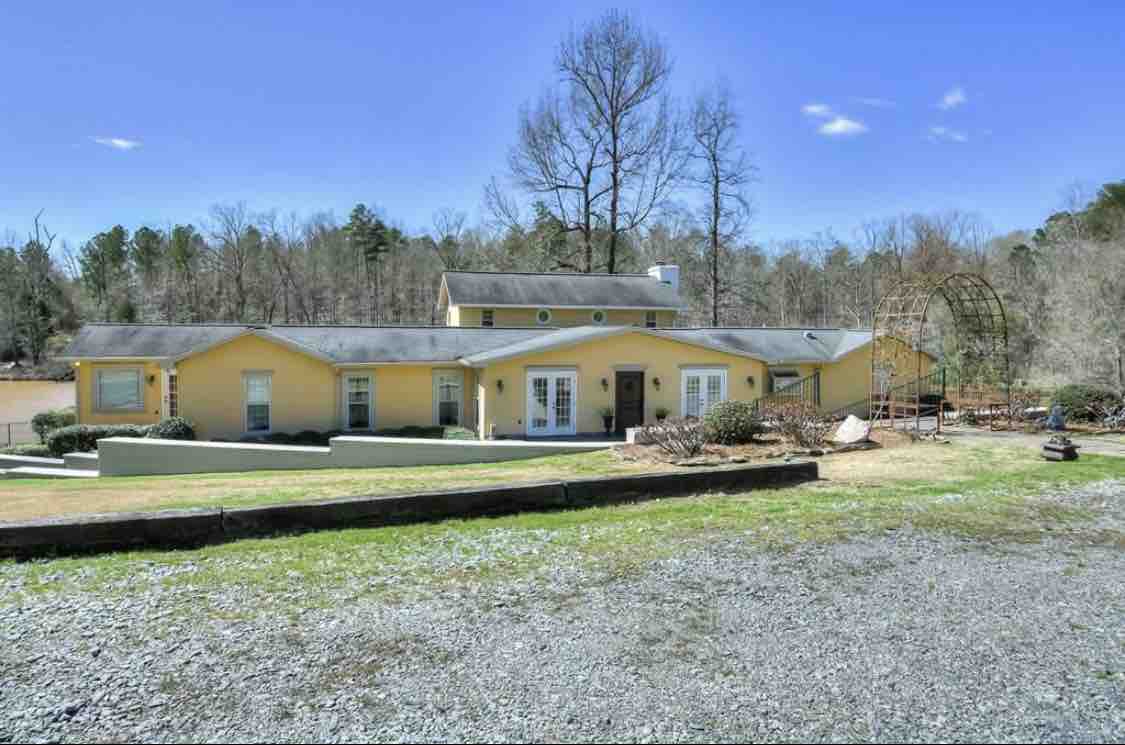 A single-story yellow house is viewed from the front, surrounded by a green lawn and bordered by gravel. Large windows and glass doors enhance natural light, while trees in the background provide a natural setting. An archway for climbing plants is visible on the right.
