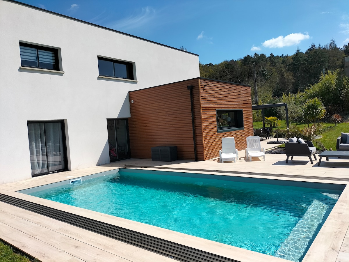 A modern house is presented with a sleek outdoor pool, surrounded by a wooden deck. Lounge chairs are positioned beside the pool, offering relaxation. The exterior features a combination of white and wooden panels, with large glass doors leading into the interior.