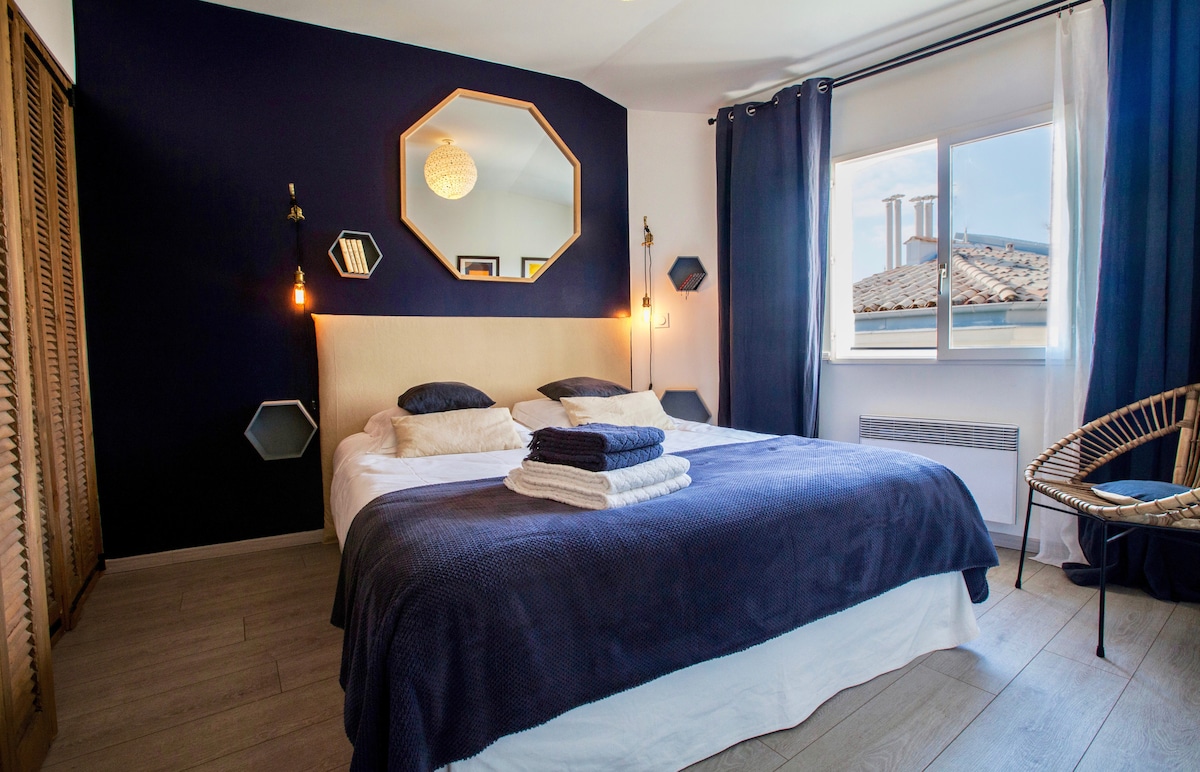 A cozy bedroom featuring a spacious bed dressed in navy linens and layered with neatly folded towels. The room is decorated with a geometric mirror, warm lighting fixtures, and stylish accents. A large window allows natural light to fill the space, showcasing views of the rooftops.