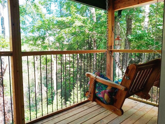 A wooden swing is positioned on a covered porch surrounded by greenery. The railings provide a view of the trees beyond, creating a serene outdoor retreat. Natural light filters through the trees, enhancing the tranquil atmosphere on the deck.