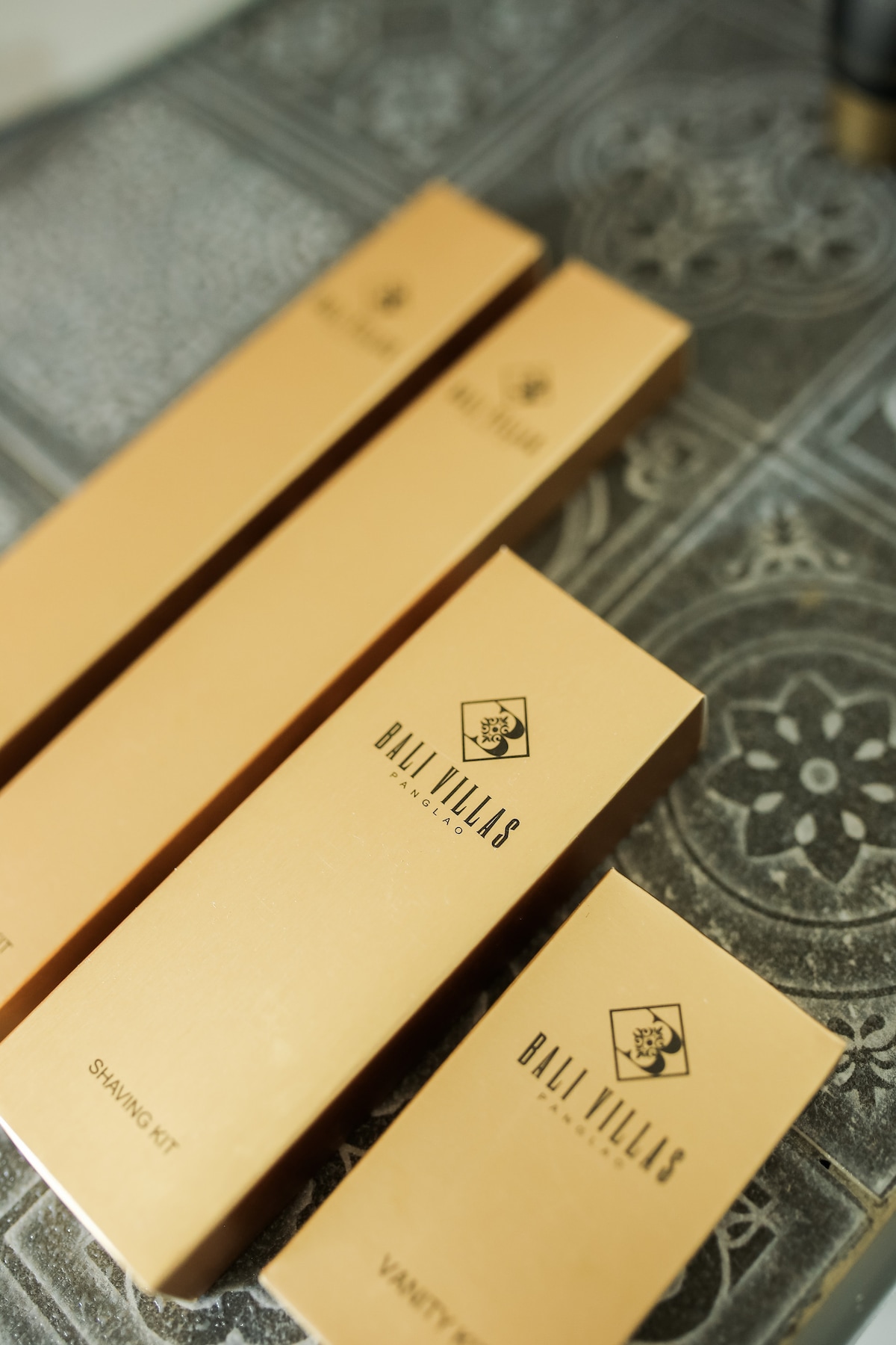 A neatly arranged collection of bath amenities includes a vanity kit, shampoo kit, and other toiletries, all presented in elegant gold packaging. The products prominently feature the Bali Villas logo, adding a touch of sophistication to the bathroom setting.