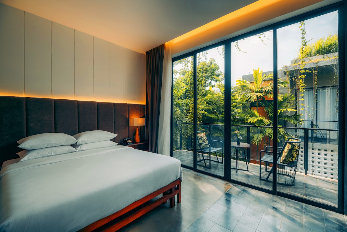 A bright guestroom features a spacious bed with crisp white linens, framed by large glass doors revealing a lush outdoor view. A well-lit seating area, complete with two chairs and a table, provides a space for relaxation, complemented by soft natural light.