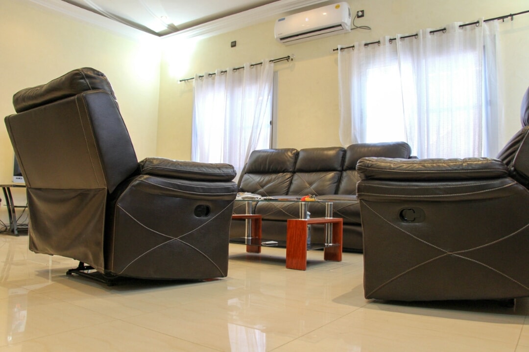 A bright and airy living room features leather recliners positioned for comfort, with a sleek coffee table at the center. Natural light filters through sheer curtains, illuminating the glossy tiled floor. A small decorative item is placed on the coffee table, adding a subtle accent.