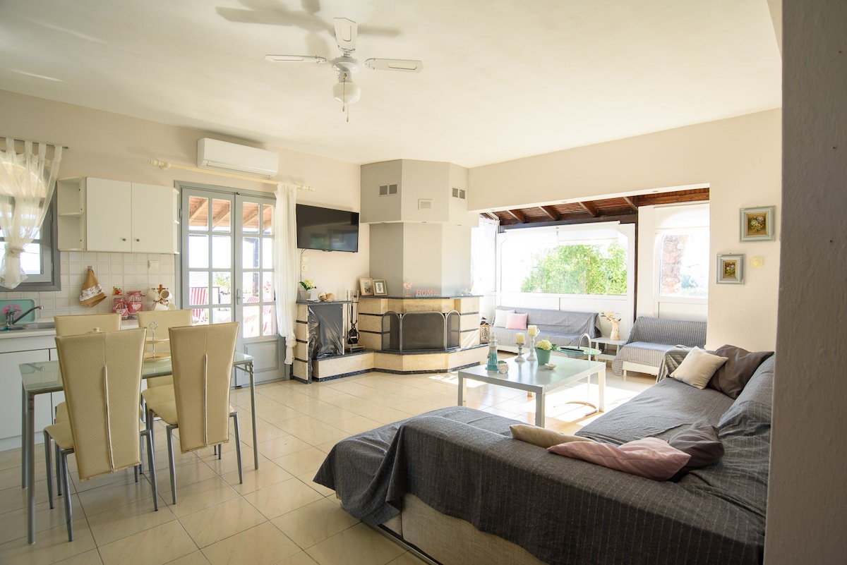 A spacious living area is equipped with a comfortable sofa and dining table, complemented by a modern television. Natural light floods the room through large windows, showcasing a cozy ambiance. The fully equipped kitchen is visible in the background, enhancing the functionality of the shared space.