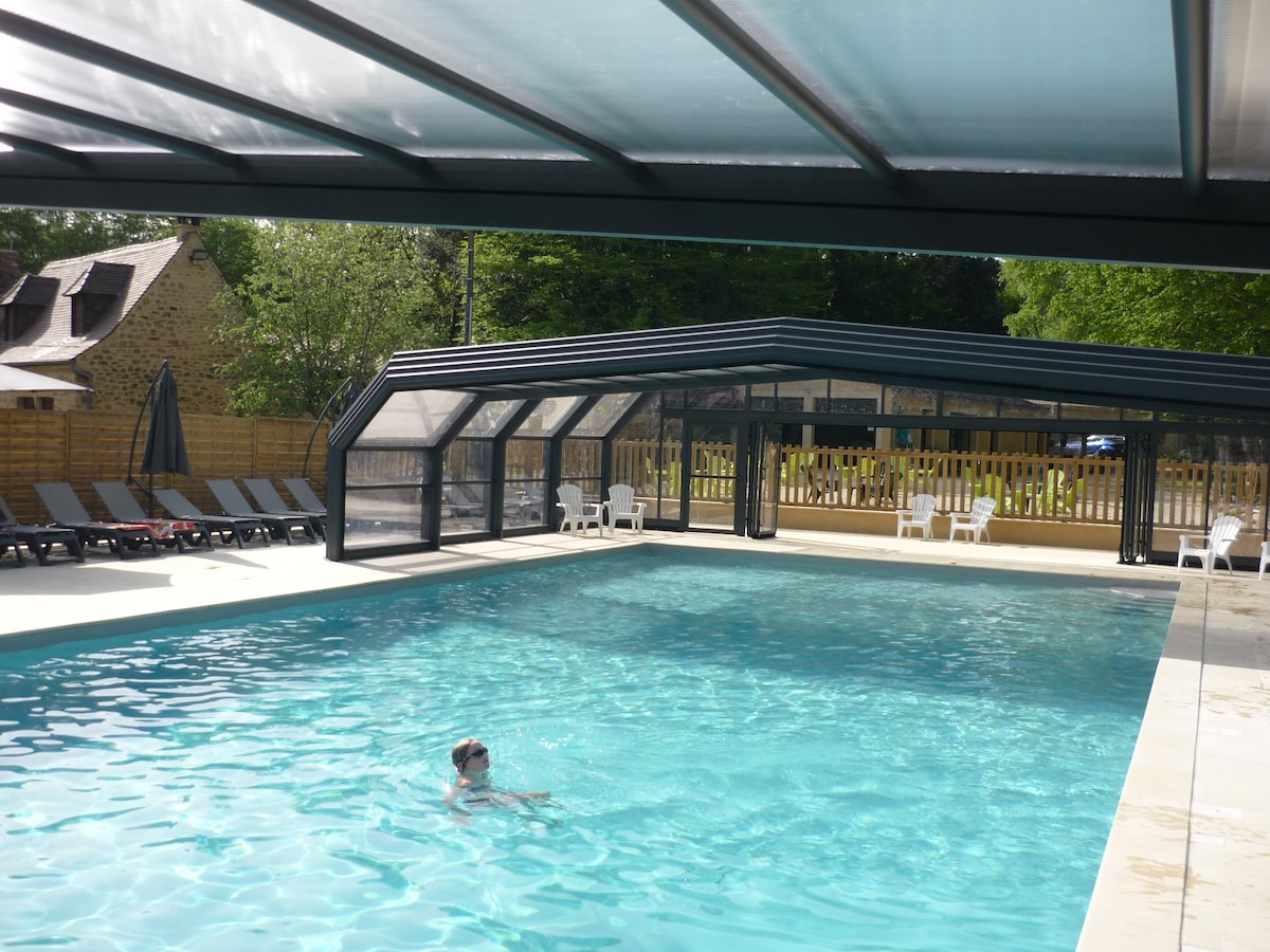 A covered heated pool occupies the center of the image, featuring clear blue water. Surrounding the pool are several lounge chairs arranged neatly under the shelter. An outdoor fence and grassy areas are visible in the background, providing a sense of openness.