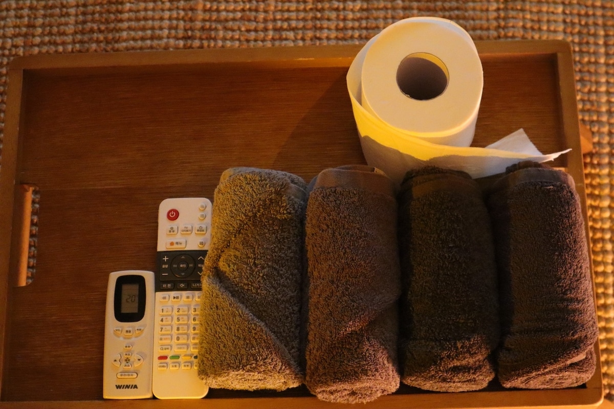 A wooden tray holds essential items including a remote control, a roll of toilet paper, and several neatly arranged dark towels. The soft lighting creates a subdued atmosphere, highlighting the textures and colors of the items.