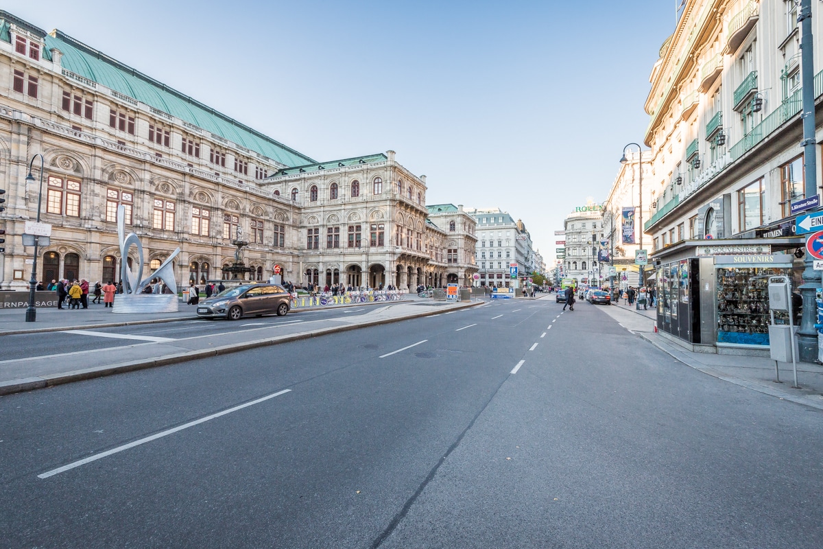A wide street in front of the Vienna State Opera is depicted, featuring a historic building with ornate architectural details and a green roof. Cars are seen driving along the road, while pedestrians are visible walking nearby. Shops and cafes line the street, enhancing the urban atmosphere.