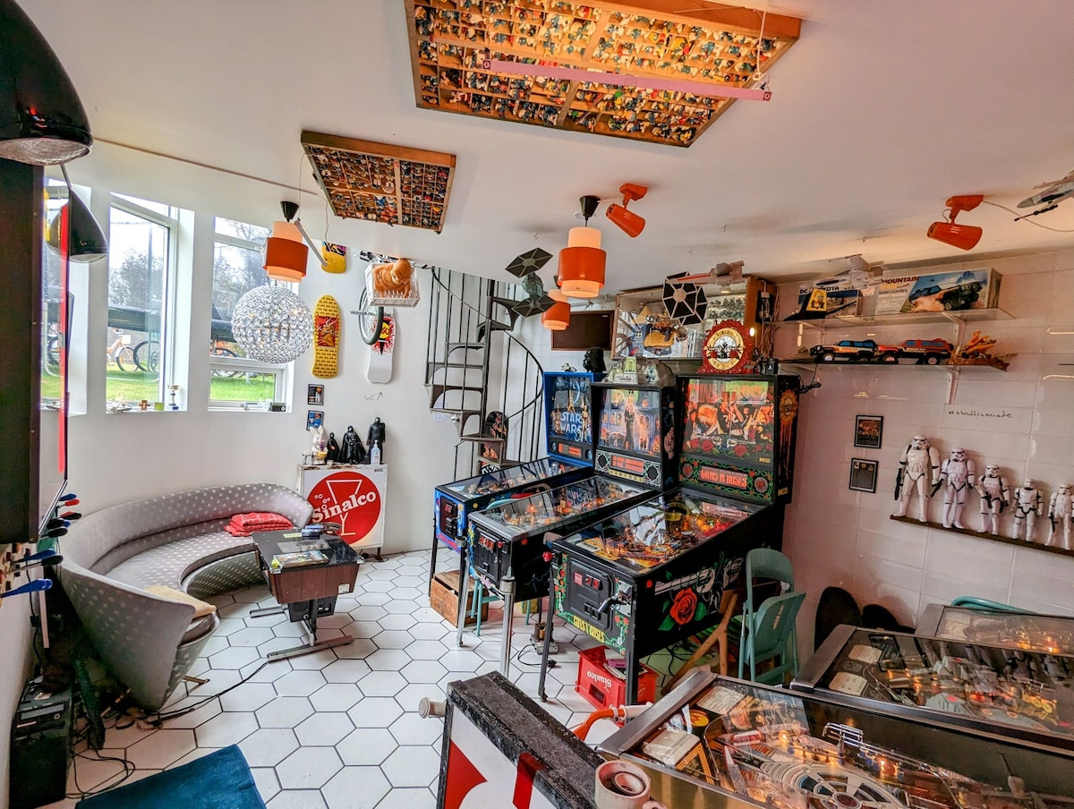 An engaging entertainment area features six pinball machines and two arcade machines, inviting exploration. Various toys and collectibles are displayed on shelves. Natural light fills the space from large windows, while unique ceiling decor adds character. A spiral staircase leads to the upper floor.