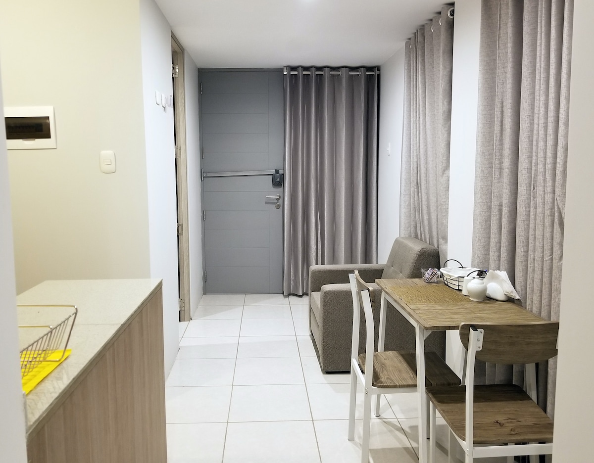 The studio apartment features a light and airy layout with a cozy seating area and a dining table set for two. Neutral-toned curtains frame the window, while a sleek kitchen area provides convenient access to cooking essentials.