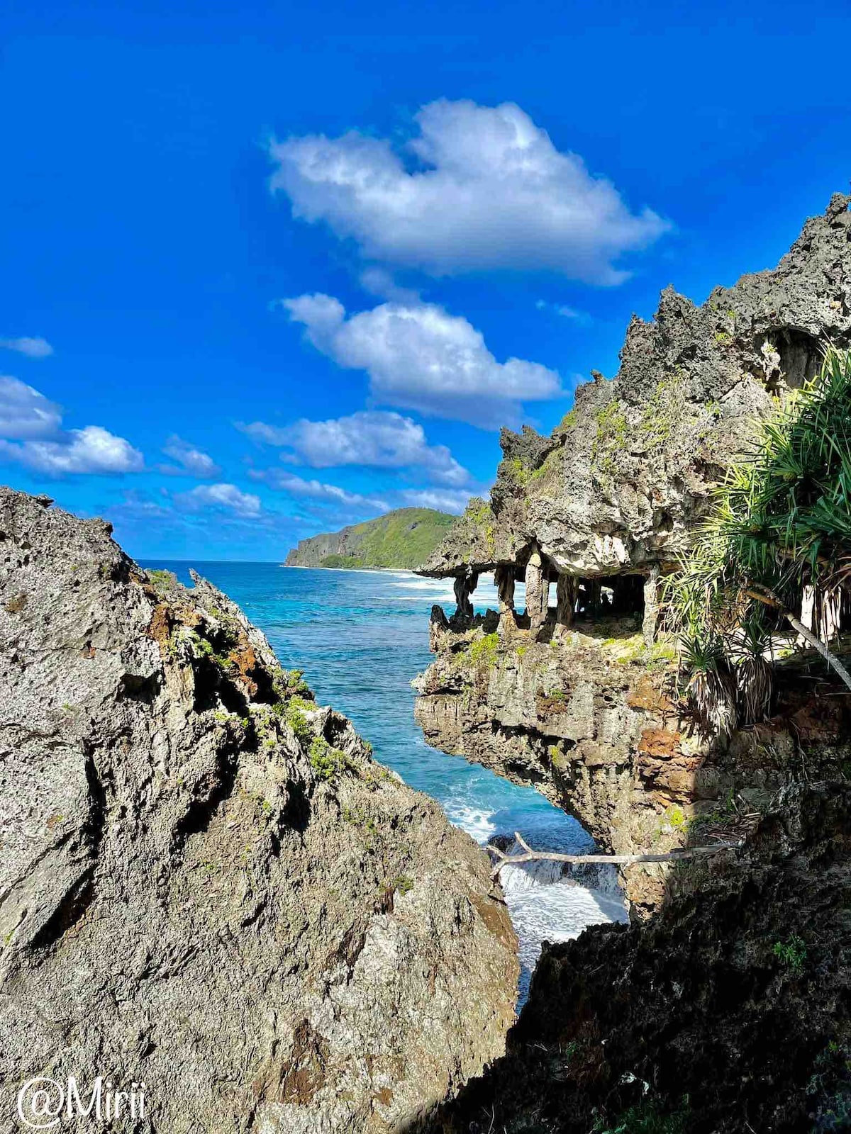 Rocky coastal formations frame a serene ocean view, with gentle waves lapping against the shore. Lush greenery is visible on the cliffs, and a bright blue sky with fluffy clouds adds to the scene's tranquility.