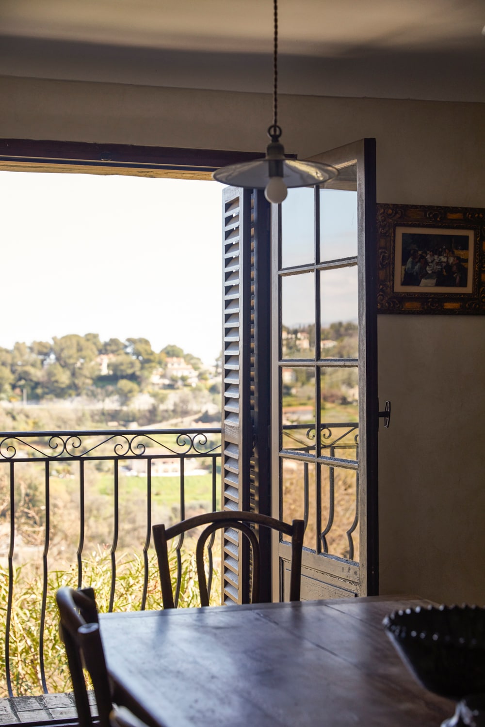 An open doorway reveals a scenic view of the landscape beyond, featuring rolling hills and greenery. A wooden dining table with simple chairs is positioned in front of the door, where natural light filters in, creating a warm ambiance.