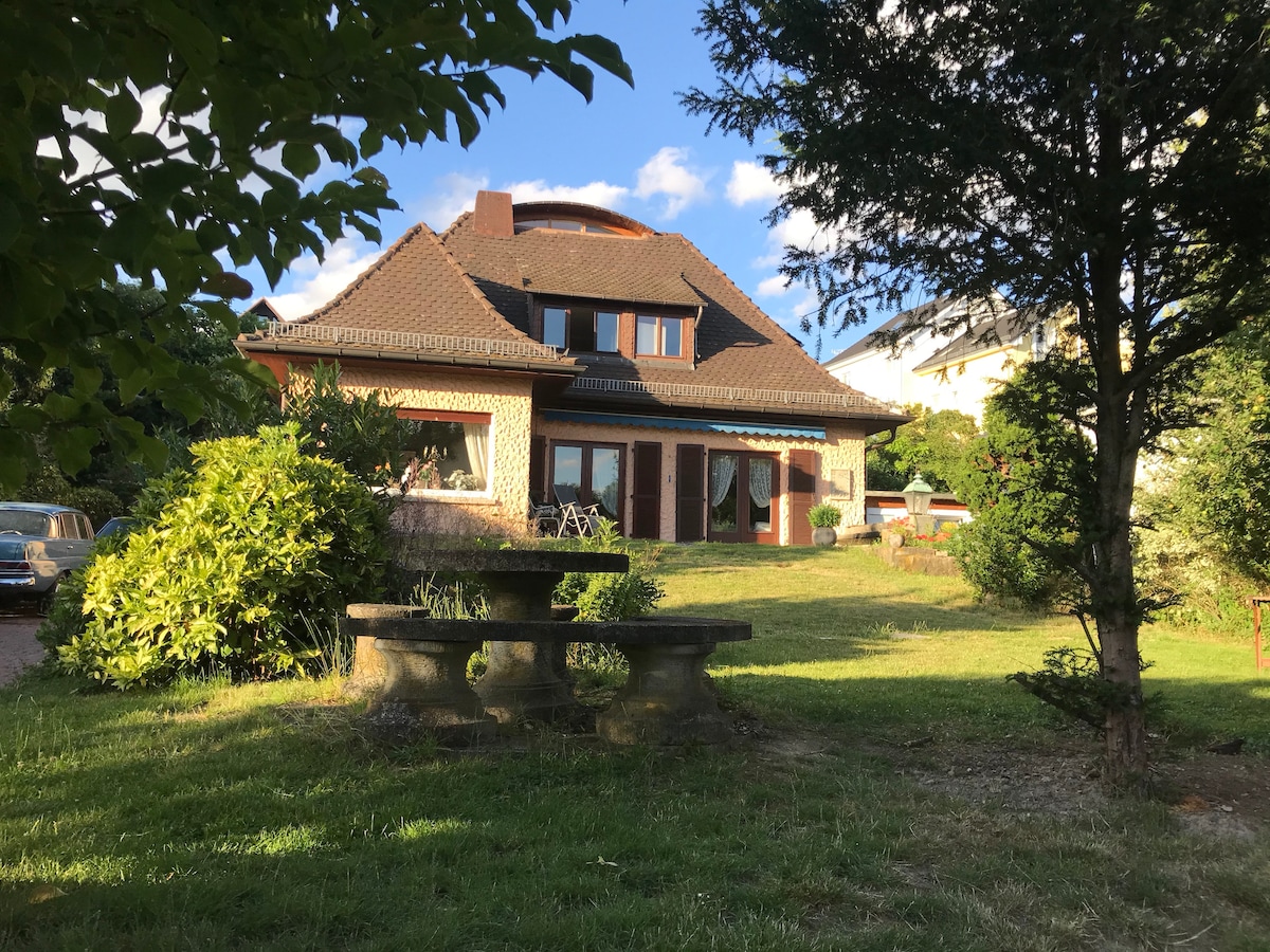A modern house with a unique roof design is surrounded by lush greenery. The exterior features large windows and a terrace that opens to the garden. A stone table with benches is positioned prominently in the foreground, amidst the well-kept lawn.
