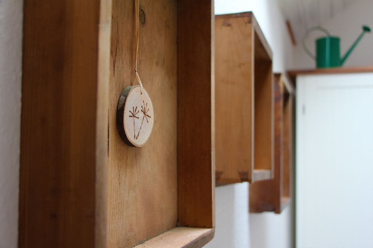 A rustic display of wooden shelves is attached to a white wall. One shelf features a wooden tag with a flower design, while the others remain empty. A green watering can is visible in the background, alongside a door leading to another room.