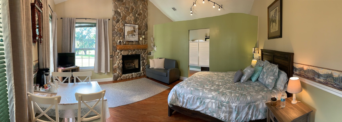A spacious room is presented, featuring a queen size bed adorned with floral bedding. A comfortable seating area includes a gray sofa beside a stone fireplace. Natural light filters through large windows, illuminating a dining table and enhancing the room's welcoming atmosphere.