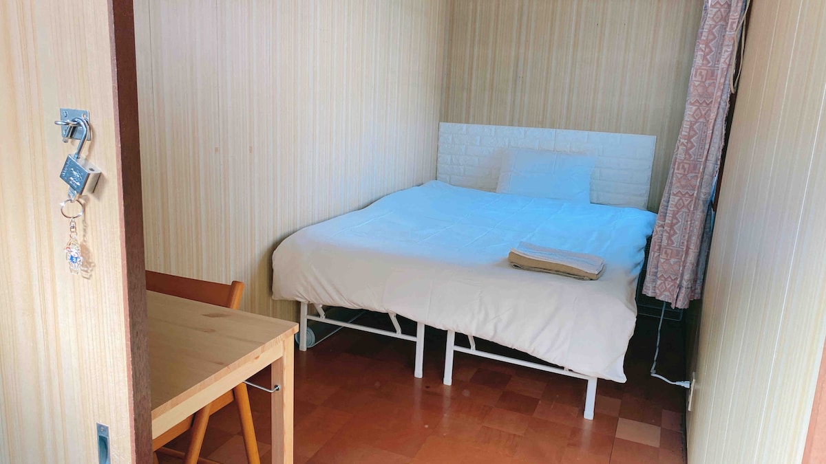 The room features a spacious bed equipped with clean bedding. A simple wooden table and chair are positioned nearby, providing a functional workspace. Natural light enters through a window partially covered by a curtain, illuminating the warm, wood-patterned flooring.