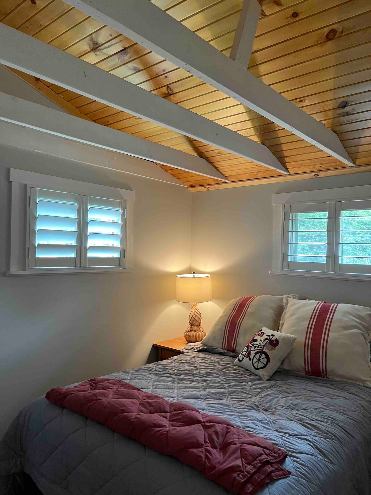 A cozy bedroom features sloped wooden ceilings and two windows adorned with plantation shutters. Soft light is emitted from a bedside lamp, illuminating a neatly made bed with decorative pillows and a pink throw at the foot.