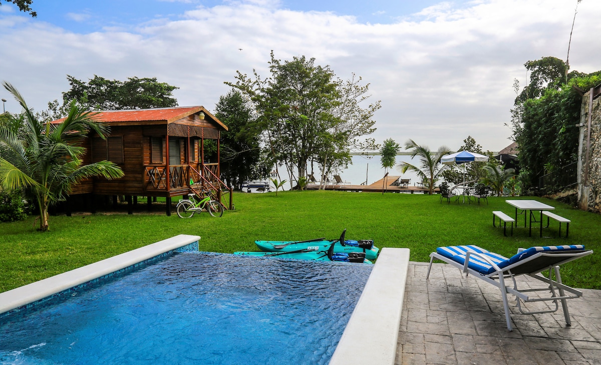A wooden bungalow is set in a lush green yard, with a refreshing swimming pool nearby. Recreational equipment, such as kayaks, is arranged beside the pool. Comfortable lounge chairs are positioned for relaxation, while views of the lagoon are visible in the background.