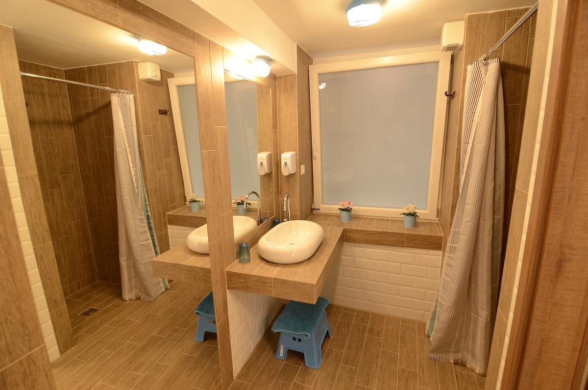 The modern bathroom features light brown tiled walls and a spacious shower area. A dual sink vanity is centered against the wall with illuminated mirrors on either side. Large windows provide natural light, complemented by small potted plants on the sill.