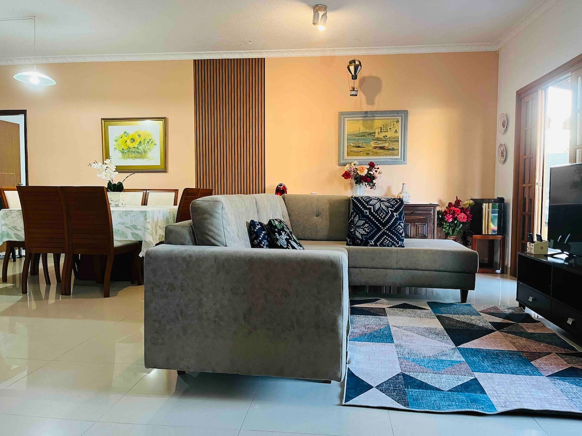 A spacious living area features a comfortable sectional sofa adorned with decorative pillows. A geometric area rug lies beneath, contrasted by polished tile flooring. In the background, a dining table is set for six, complemented by wall art and floral arrangements.