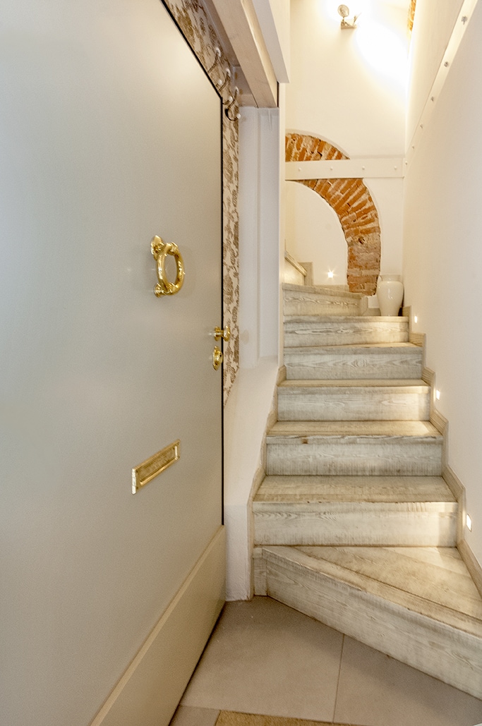 An entrance door with a brass knocker leads to a softly lit staircase made of stone, featuring subtle lighting along the steps. Exposed brick walls add texture, creating a warm and inviting entryway.