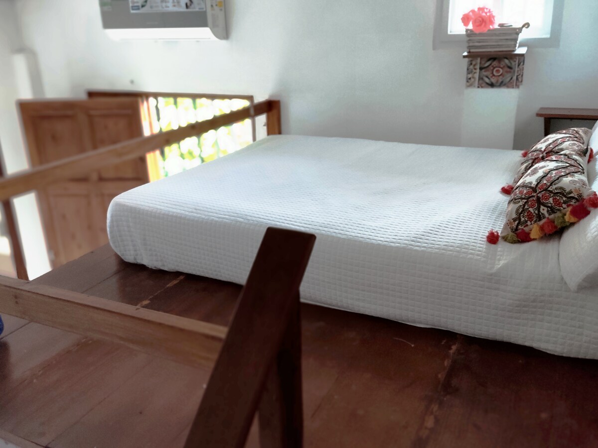 A spacious loft area is presented, featuring a neatly made bed with decorative pillows. Natural light filters through a nearby window, illuminating the light-colored walls and wooden structure. The air conditioning unit is visible, ensuring comfort in the space.