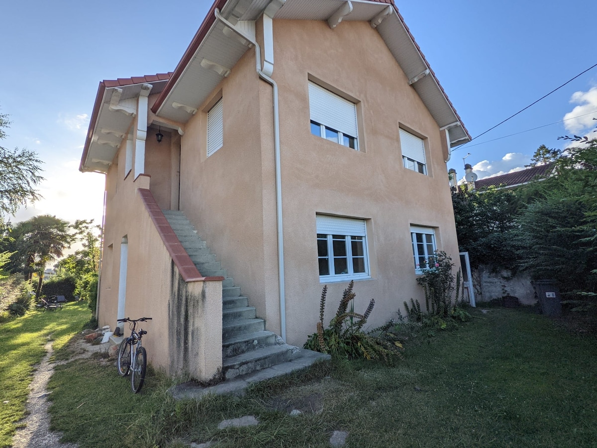 A two-story house is depicted, featuring a light-colored exterior with several windows. The image shows a staircase leading to the entrance, surrounded by a grassy area and shrubs. A bicycle is parked nearby, contributing to the outdoor setting.