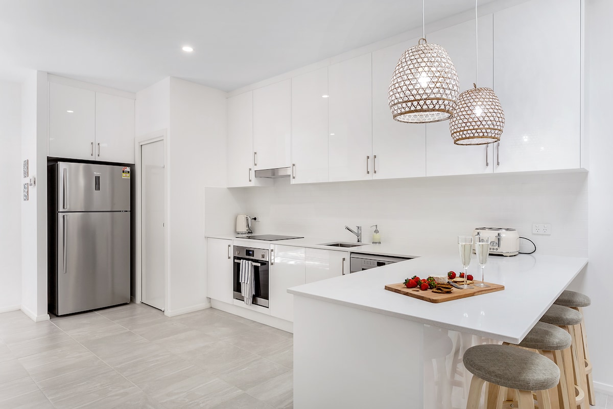 A modern kitchen features sleek white cabinetry and stainless-steel appliances, including a large refrigerator and a built-in oven. A central island with a light wood finish is accompanied by three stools. Two pendant lights hang above, adding a soft glow to the clean, open space.