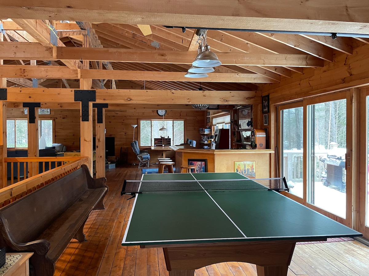An open interior space showcases a green ping pong table centered amidst wooden beams and a warm finish. Large windows allow natural light to fill the room, offering views of the surrounding trees. A wooden bench and seating area are visible in the background.