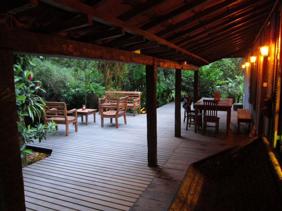 A spacious wooden deck is illuminated by soft lighting, surrounded by lush greenery. Several wooden chairs and a dining table create a welcoming space for relaxation. The natural materials blend harmoniously with the surrounding environment, inviting guests to enjoy the tranquility of the setting.