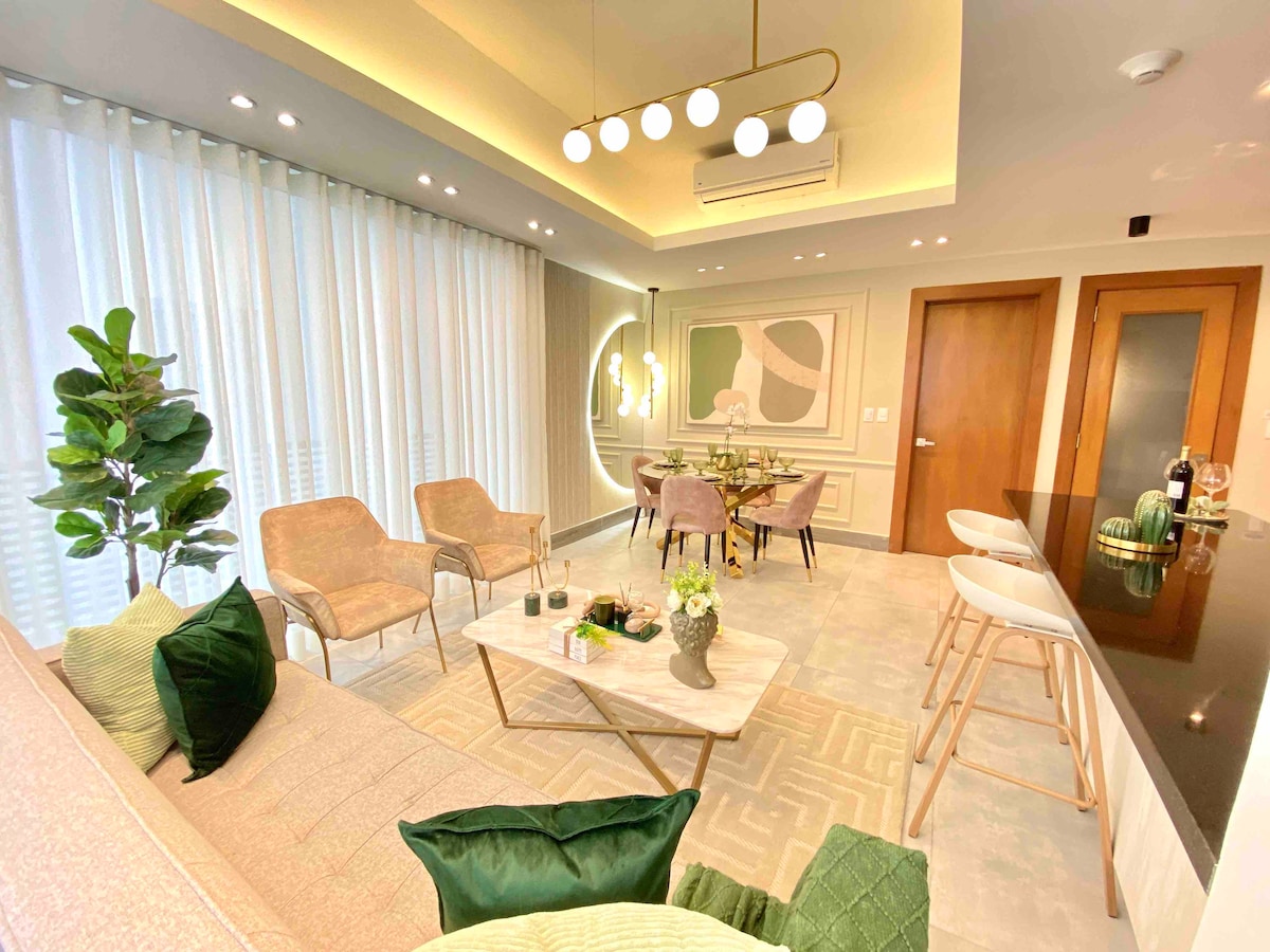 A modern living and dining area features soft seating with throw pillows, a sleek dining table, and elegant decor. Natural light filters through sheer drapes, highlighting the contemporary design elements, including a minimalist chandelier and a potted plant that adds a touch of greenery.