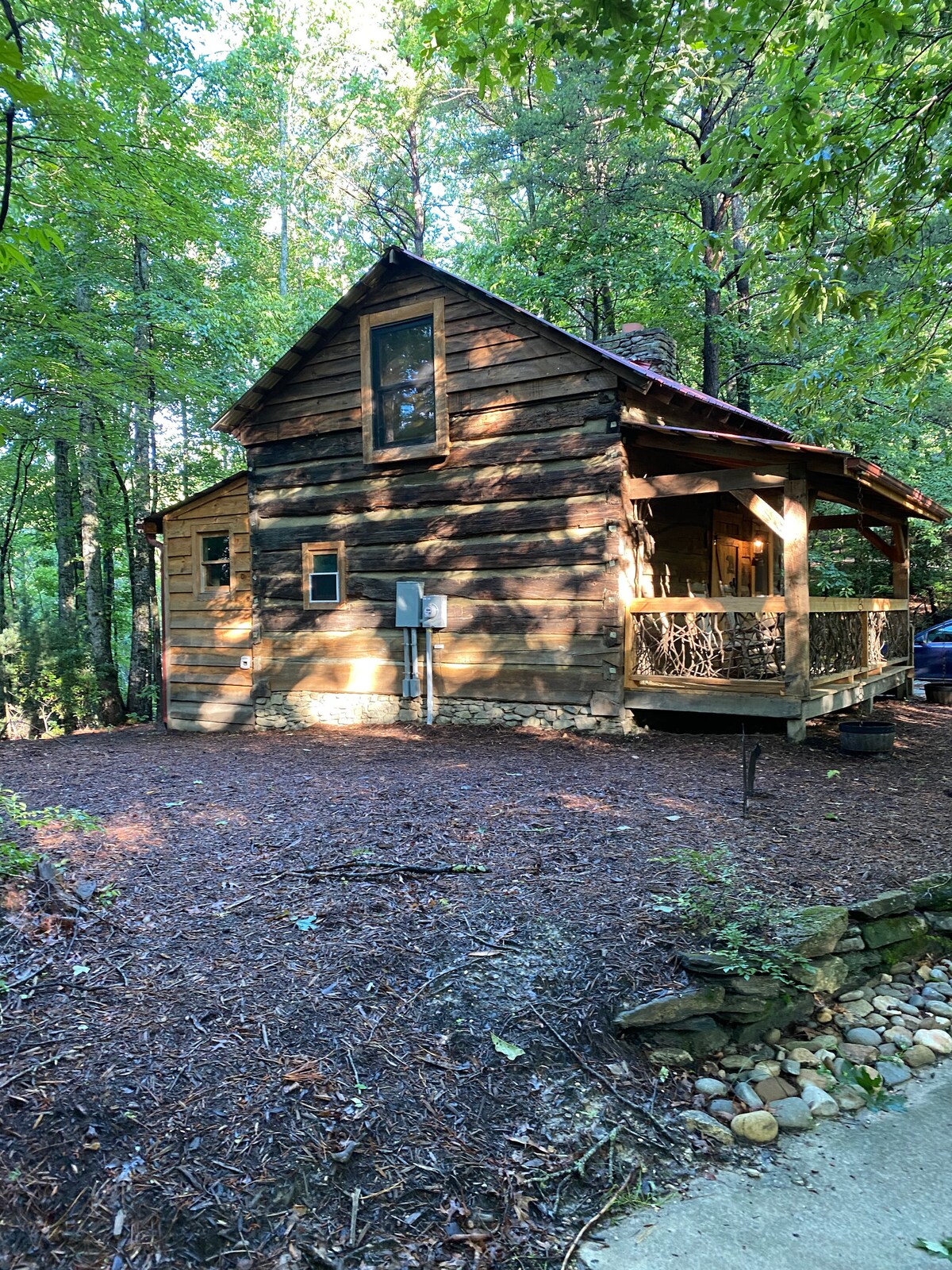 A rustic cabin with a wooden exterior is surrounded by tall trees, providing a serene and secluded environment. The front porch features a simple railing, and a window is visible on the upper level. The natural landscape is enhanced by a layer of mulch and small stones along the path.